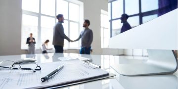 Personas de fondo estrechando la mano y en primer plano un contrato y un lapicero sobre este.